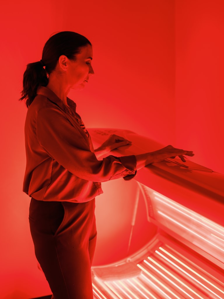 Woman standing beside a red-lit tanning bed, touching its cover.