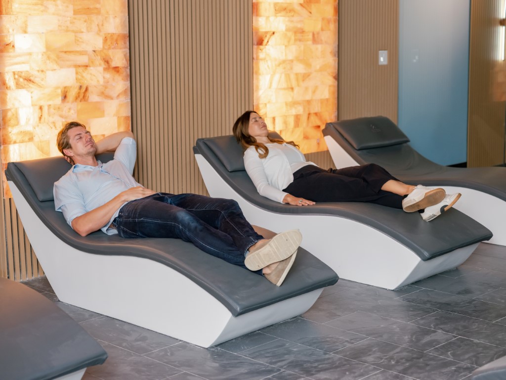 Two adults recline on modern curved lounge chairs in a spa-like lounge area, relaxing with eyes closed.