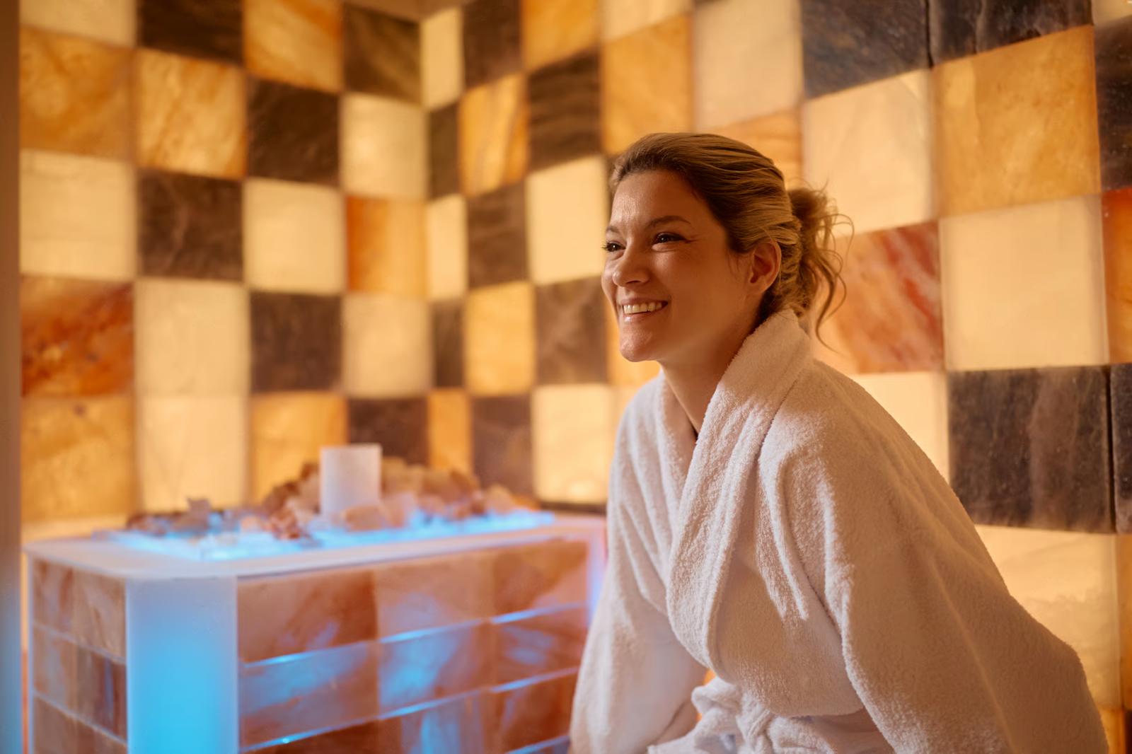 wmremove-transformed - 2026-10-09T145513212 - Scape Relaxation Smiling woman in a white bathrobe seated in a warm, tiled spa room with blue-lit water feature behind her.